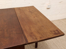Load image into Gallery viewer, A close-up of a dark brown wooden table, showing the wood grain and a visible seam on the tabletop, set against a white brick wall and beige carpet.

