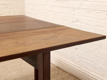 Load image into Gallery viewer, Close-up of a dark brown wooden table corner with a white painted brick wall and light beige rug in the background.
