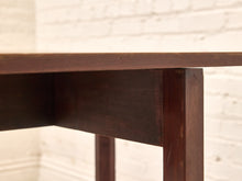 Load image into Gallery viewer, Close-up of a dark brown wooden table showing the tabletop edge, leg joint, and part of the leg, set against a softly blurred white brick wall background.
