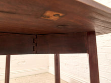Load image into Gallery viewer, Close-up underside view of a dark brown wooden table, showing the joint connection and leg structure in a room with a white brick wall and carpeted floor.
