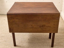 Load image into Gallery viewer, A mid-century modern wooden table with a drop-leaf design, featuring a warm medium-brown wood finish and dark legs, set on a textured beige rug against a white brick wall.
