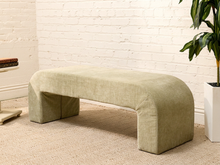Load image into Gallery viewer, An upholstered bench with a curved top and blocky legs in a light sage green fabric, set against a white brick wall on a beige carpet with a potted plant nearby.

