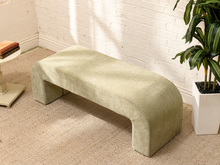 Load image into Gallery viewer, An upholstered bench in a light sage green fabric, styled in a bright room with a white brick wall, beige carpet, a potted plant, and a small side table.
