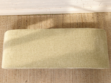 Load image into Gallery viewer, A rectangular upholstered bench cushion in a light beige textured fabric, shown from a top-down view on a woven jute rug against a white brick wall.
