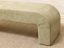 Load image into Gallery viewer, A close-up of a curved upholstered bench or ottoman in a light sage green fabric, shown on a woven rug against a white brick wall.
