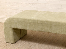 Load image into Gallery viewer, A modern upholstered bench with a curved front and blocky legs, covered in a textured sage green fabric, set on a beige carpet against a white brick wall.
