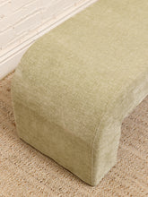 Load image into Gallery viewer, A close-up of a light sage green upholstered bench corner, showing the textured fabric, set on a woven jute rug against a white brick wall.
