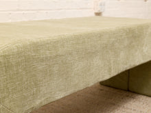 Load image into Gallery viewer, A close-up of a light olive green upholstered bench or ottoman with a textured woven fabric, shown in a room setting with a white brick wall and carpeted floor.

