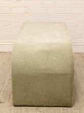 Load image into Gallery viewer, A rectangular upholstered ottoman or bench with rounded top corners, covered in a textured light sage green fabric, set on a woven rug against a white brick wall.
