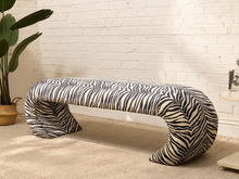 Load image into Gallery viewer, A sculptural curved bench upholstered in a black and cream zebra-print fabric, set against a white brick wall and beige carpet with a potted plant on the left.
