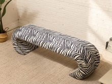 Load image into Gallery viewer, A modern curved bench upholstered in a black and off-white zebra print fabric, set against a white brick wall and beige carpet.
