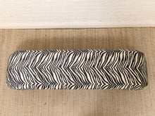 Load image into Gallery viewer, A rectangular upholstered bench with a bold black and off-white zebra stripe pattern, resting on a textured beige rug against a white brick wall.
