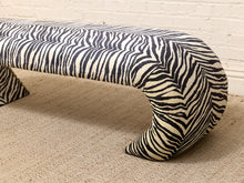 Load image into Gallery viewer, A modern curved bench upholstered in a bold black and off-white zebra print fabric, set against a light beige textured rug and a white brick wall.
