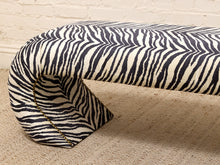 Load image into Gallery viewer, A curved upholstered bench with a bold black and off-white zebra stripe pattern, resting on a beige textured rug against a white brick wall.
