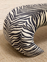 Load image into Gallery viewer, A curved upholstered bench or stool with a bold black and off-white zebra stripe pattern, shown on a textured beige rug against a white brick wall.
