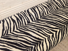 Load image into Gallery viewer, A close-up of a zebra-patterned fabric or wallpaper roll, showing bold black and cream stripes in a chevron arrangement, positioned over a white brick baseboard and light-colored floor.
