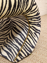 Load image into Gallery viewer, Close-up of a zebra-print upholstered chair or cushion with black and cream stripes, a curved arm, and a gold beaded trim detail on a woven beige floor against a white brick wall.

