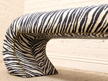 Load image into Gallery viewer, A curved upholstered bench with a bold black and off-white zebra print, featuring gold contrast stitching along the edge, set against a cream brick wall and light carpet.
