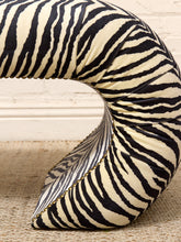 Load image into Gallery viewer, A curved, zebra-print upholstered cushion or pet bed with black and cream stripes, resting on a woven rug against a white brick wall.
