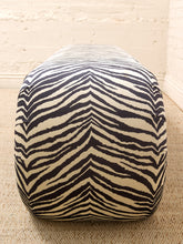Load image into Gallery viewer, A low, rounded pouf ottoman with a bold black and cream zebra stripe pattern, set on a textured beige rug against a light beige brick wall.
