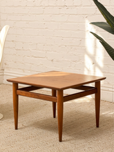 Load image into Gallery viewer, A square wooden side table with a warm medium-brown finish, featuring a lower support rail and tapered legs, set on a textured beige rug against a white brick wall.
