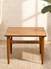 Load image into Gallery viewer, A square wooden side table with a warm medium-brown finish, featuring tapered legs and a lower support rail, set on a textured beige rug against a white brick wall.
