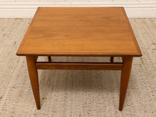 Load image into Gallery viewer, A mid-century modern square wooden coffee table with a warm medium-brown finish, featuring a lower support rail and tapered legs, set on a textured beige rug.
