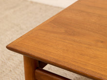 Load image into Gallery viewer, Close-up of a mid-century modern wooden table corner with a warm medium-brown finish and visible wood grain, set against a light beige carpet.
