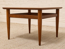 Load image into Gallery viewer, A mid-century modern wooden side table with tapered legs and a lower shelf, shown on a textured beige rug against a white brick wall.
