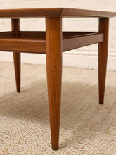 Load image into Gallery viewer, Close-up of a mid-century modern wooden table leg and apron, showing a warm medium-brown wood finish, set on a textured beige woven rug against a white brick wall.
