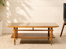 Load image into Gallery viewer, A mid-century modern wooden coffee table with a glass top and lower shelf, set against a white brick wall with leaf shadows.
