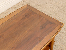 Load image into Gallery viewer, A close-up of a wooden side table with a warm medium-brown wood grain finish and a clear glass top, set on a beige textured carpet.
