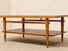 Load image into Gallery viewer, A mid-century modern wooden coffee table with a glass top, two wooden shelves, and brass legs, set against a white brick wall and beige carpet.
