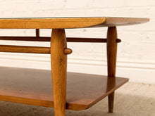 Load image into Gallery viewer, A close-up of a mid-century modern wooden coffee table with a glass top, featuring tapered legs, horizontal support bars, and a lower shelf, set against a white brick wall background.
