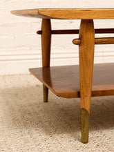 Load image into Gallery viewer, Close-up of a mid-century modern wooden coffee table with a glass top, warm medium-brown wood legs, and brass-colored metal feet, set on a textured beige rug against a white brick wall.
