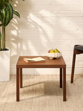 Load image into Gallery viewer, A square wooden side table with a warm medium-brown finish, styled in a bright room with a white brick wall, a potted plant, and a chair. On the tabletop sits an open book and a white scalloped bowl of fruit.
