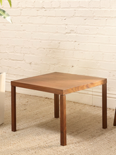 Load image into Gallery viewer, A square wooden side table with four legs, shown in a room setting against a white brick wall and beige carpet.
