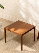 Load image into Gallery viewer, A square wooden side table with four legs, shown in a styled room setting with a white brick wall, beige rug, and soft leaf shadows.
