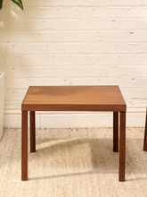 Load image into Gallery viewer, A square wooden side table with a medium brown finish and four legs, positioned on a textured beige rug against a white brick wall.

