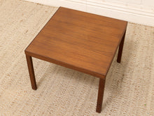 Load image into Gallery viewer, A square wooden side table with a warm medium-brown finish and visible wood grain, positioned on a textured beige rug against a white wall.
