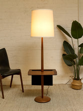 Load image into Gallery viewer, A mid-century modern floor lamp with a warm white fabric drum shade, a vertical wooden stem, and a square wooden base that doubles as a side table, shown in a styled room setting with a brick wall, chair, and potted plant.

