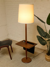 Load image into Gallery viewer, A mid-century modern floor lamp with a warm glowing white drum shade, a slender wooden stand, and a square wooden side table integrated into the base, set in a room corner with a white brick wall, beige rug, and a potted plant.
