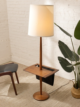 Load image into Gallery viewer, A mid-century modern floor lamp with a warm white drum shade, a medium brown wooden stand, and a circular wooden base, styled in a room corner with a white brick wall, beige carpet, and a potted plant.
