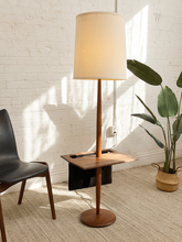 Load image into Gallery viewer, A mid-century modern floor lamp with a tall beige fabric drum shade, a wooden pole, and a square wooden side table attached at the base, styled in a bright room with a white brick wall, a black chair, and a potted plant.
