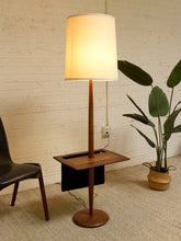 Load image into Gallery viewer, A mid-century modern floor lamp with a warm glowing white drum shade, a slender wooden pole, and a square wooden side table base, set in a room with a white brick wall, beige carpet, a black chair, and a potted plant.
