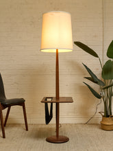 Load image into Gallery viewer, A mid-century modern floor lamp with a warm white fabric drum shade, a dark brown wooden stand, and a matching round base, styled in a room with a brick wall, a chair, and a potted plant.

