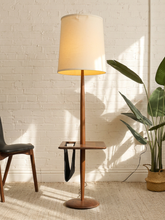 Load image into Gallery viewer, A mid-century modern floor lamp with a warm wood finish and a beige fabric drum shade, styled in a room with a white brick wall, a potted plant, and a chair.
