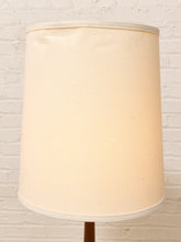 Load image into Gallery viewer, A large cylindrical fabric lampshade in a warm cream or off-white color, shown on a lamp stand against a white painted brick wall background.

