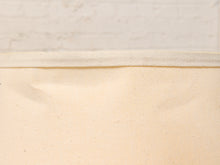Load image into Gallery viewer, A close-up of a light cream canvas fabric with a visible woven texture and a matching fabric band along the top edge, set against a white brick wall background.
