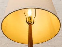 Load image into Gallery viewer, A close-up of a floor lamp with a large beige fabric drum shade, a warm yellow-gold interior, a wooden stem, and a glowing light bulb in a brass socket, set against a white painted brick wall.
